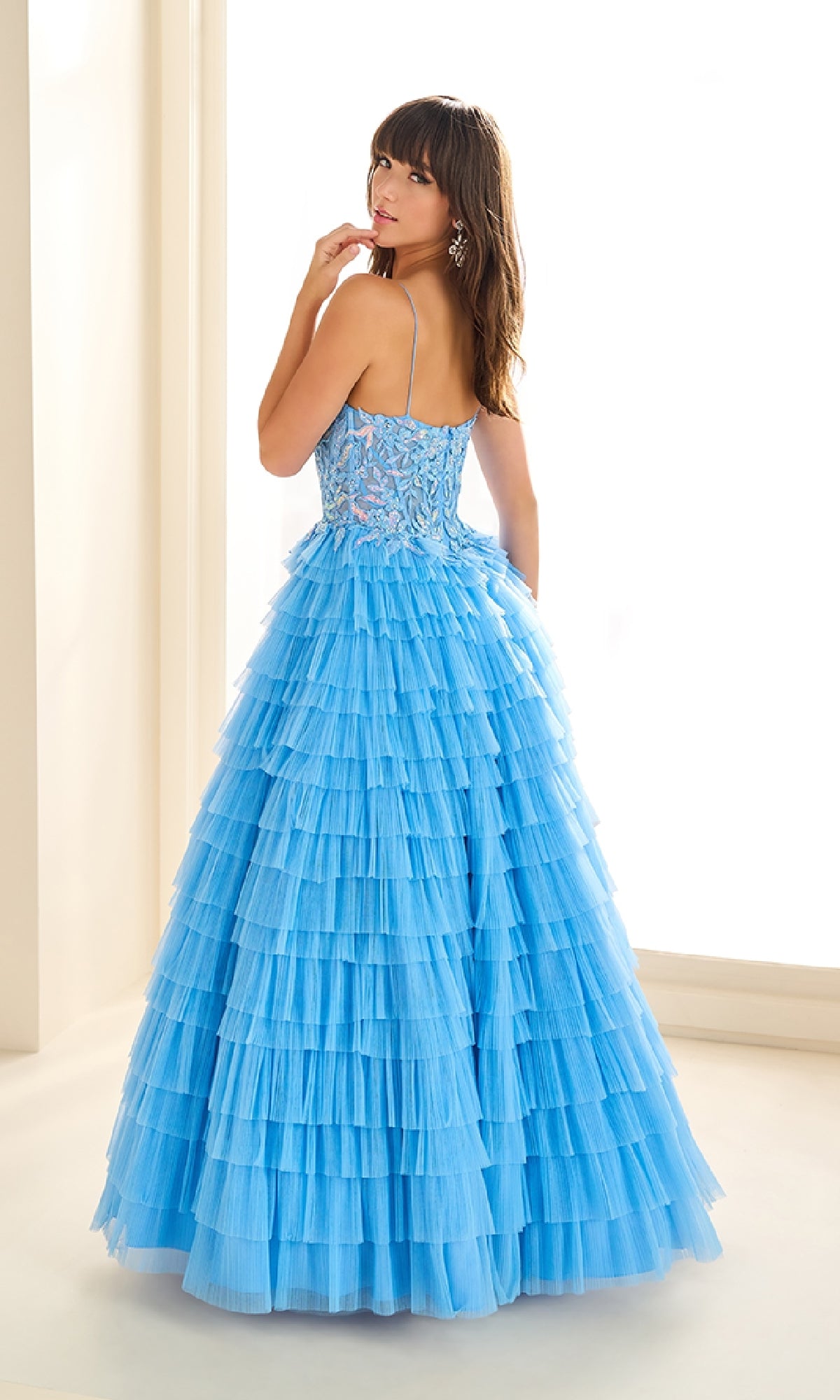 A woman stands in a bright room wearing the Ellie Wilde EW36086, a long prom dress with thin straps, a floral lace corset bodice, and light blue ruffled details.